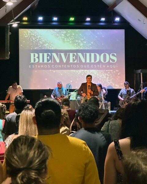 A pastor and worship team stand on stage with a sign that says "Welcome" in Spanish. 